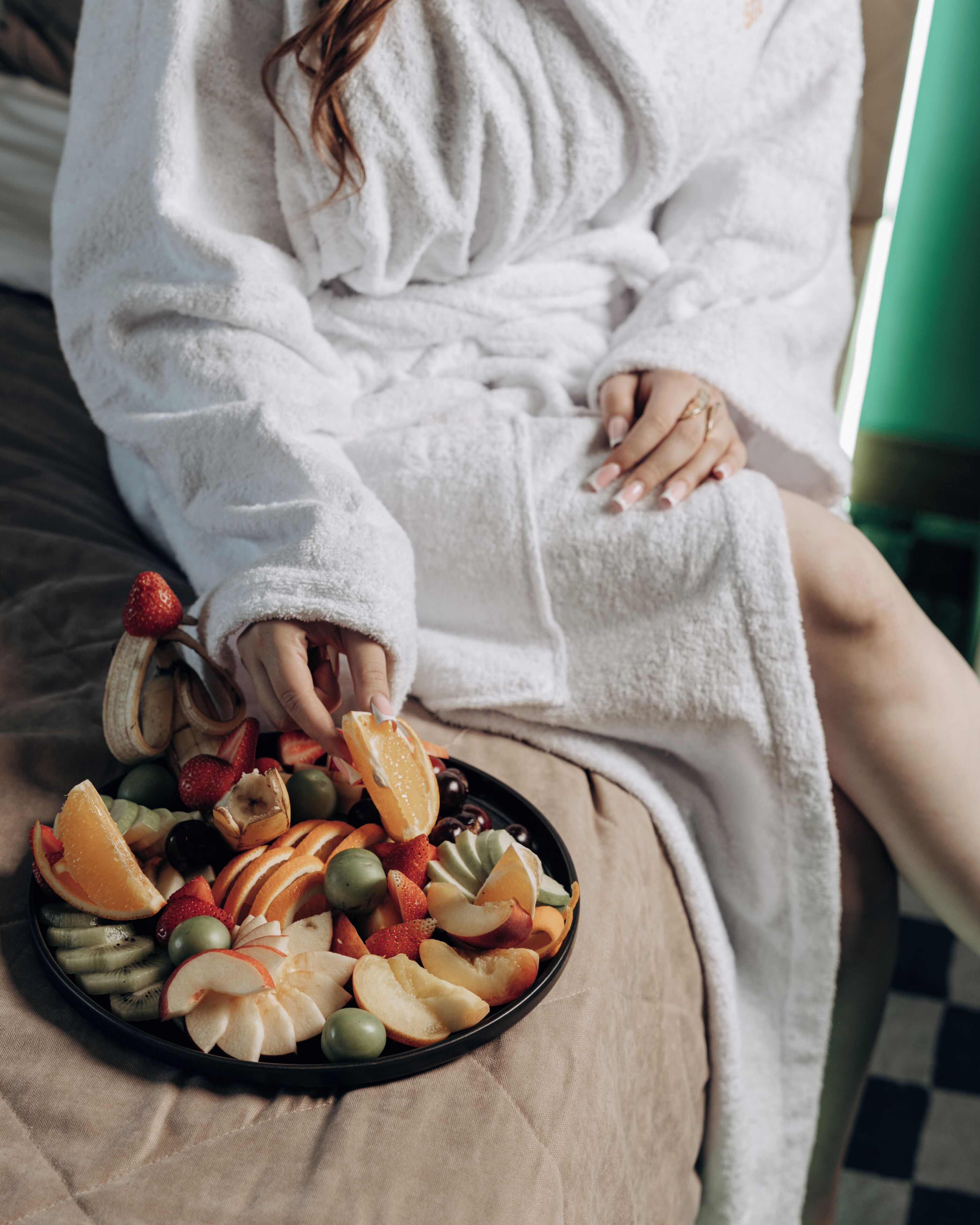 A person in a white robe sitting on a bed with a fruit platter.