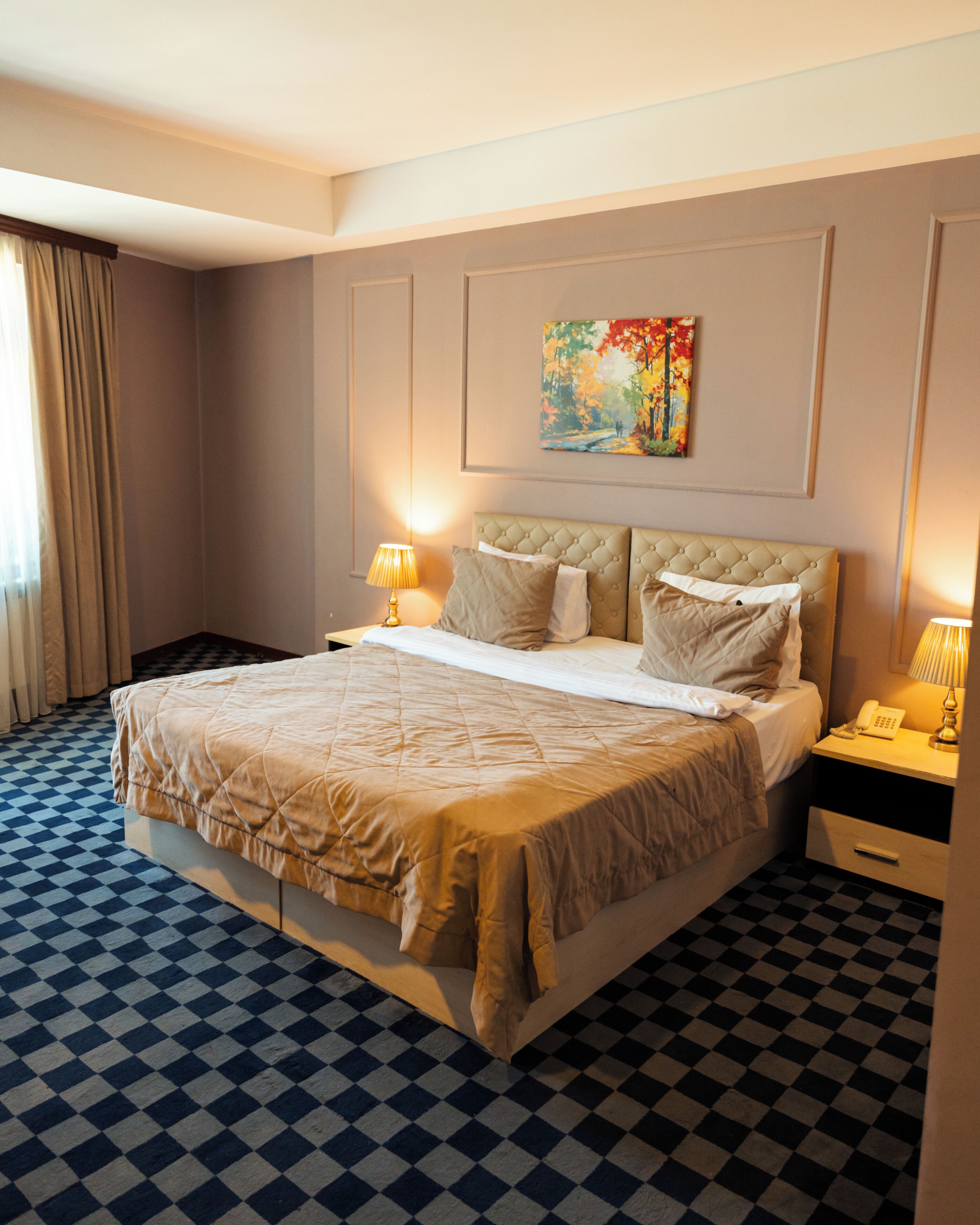 Quarto de hotel com chão xadrez, cama com cabeceira acolchoada, duas mesas de cabeceira com luminárias e um quadro na parede.