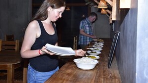 Une femme sert des plats à une table en bois, où se trouvent plusieurs assiettes.