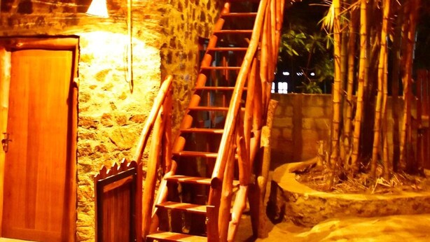 Un escalier en bois mène à un bâtiment dont le toit est fait de bambou, avec des murs en pierre et une porte en bois.