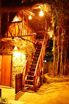 Una escalera de madera que lleva a un edificio con techo de bambú, paredes de piedra y una puerta de madera.