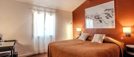 A bedroom with a large bed, two bedside lamps, a window with curtains, a desk, and a wall-mounted artwork.