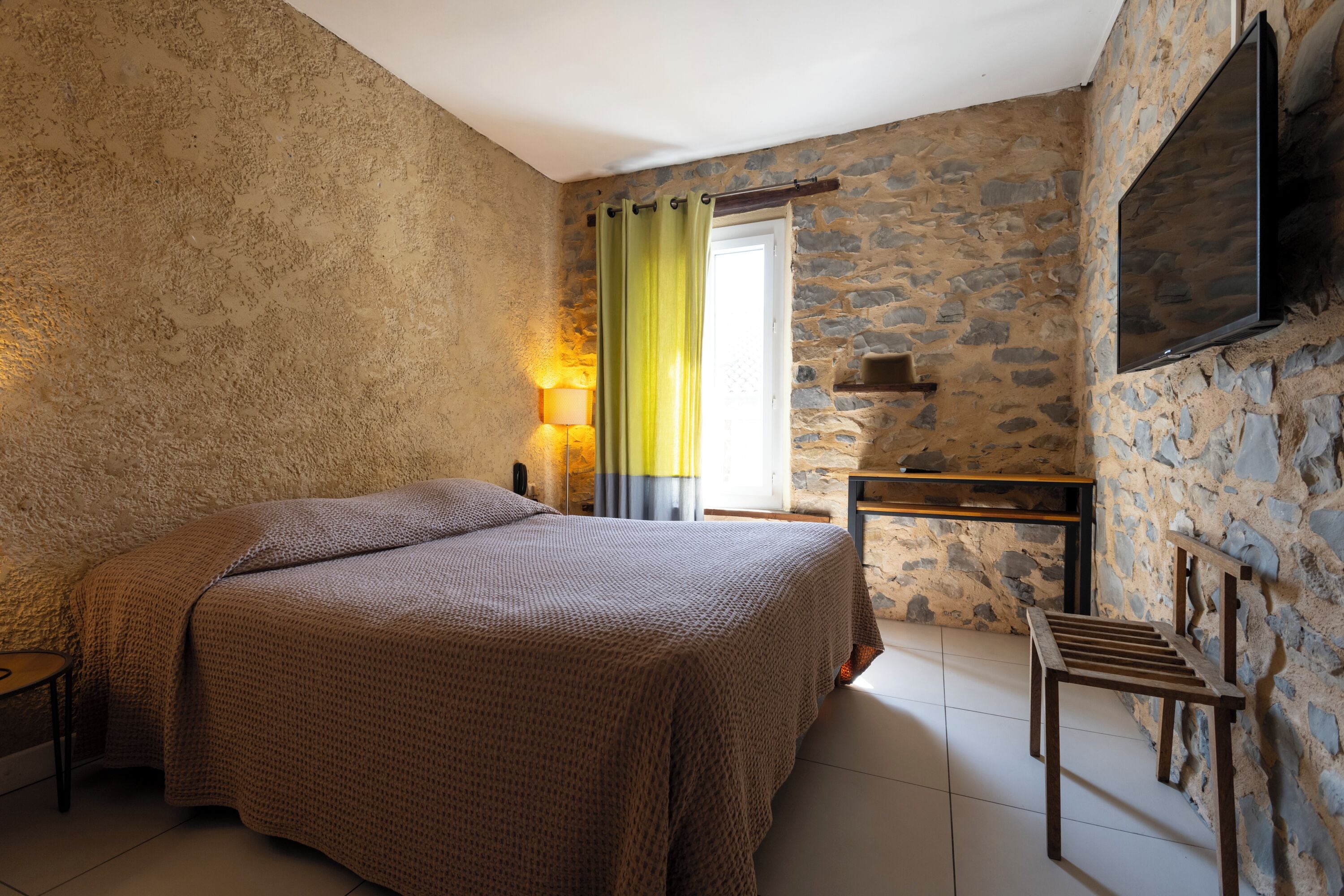 Un dormitorio con una cama grande, un escritorio, una silla, un televisor montado en la pared y una ventana con cortinas.