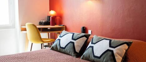 A hotel room with a bed, two pillows, a desk with a chair, a lamp, and a wall-mounted light fixture.