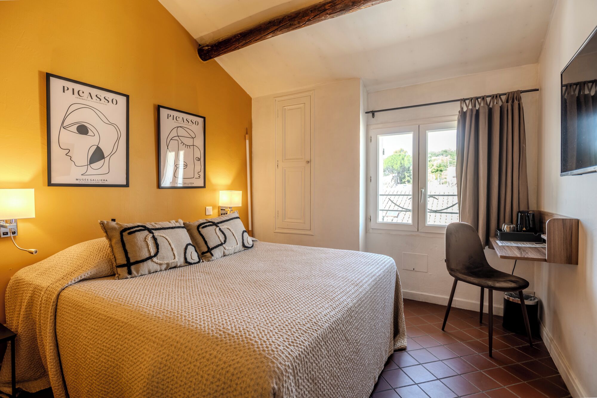 A hotel room with a bed, two wall-mounted lamps, a desk with a chair, a window with curtains, and two framed artworks on the wall.