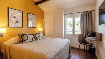A hotel room with a bed, two wall-mounted lamps, a desk with a chair, a window with curtains, and two framed artworks on the wall.