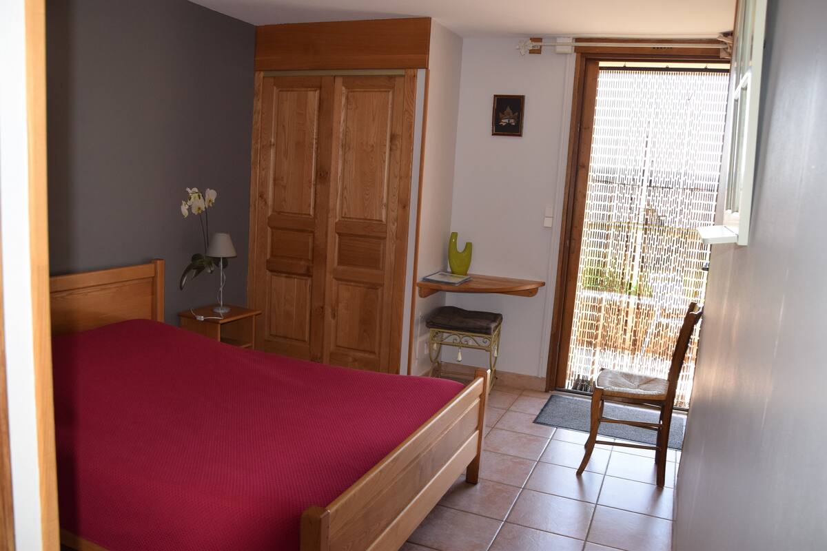 A bedroom with a wooden bed, a nightstand, a chair, a small table with a lamp, and a window with a view.