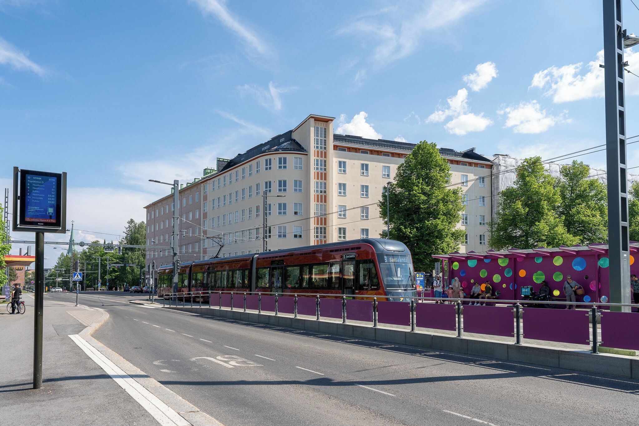 A tram station with a modern tram, a purple barrier, and a multi-story building in the background.