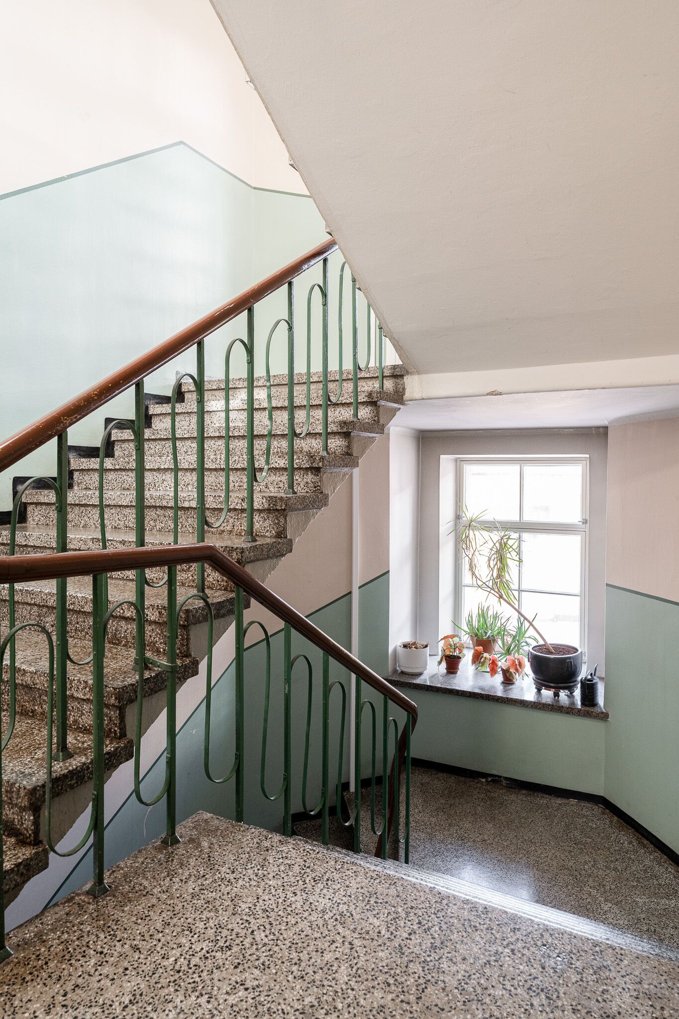 Una escalera con barandillas de metal verde y piso de granito, que conduce a una habitación con ventana y plantas en macetas.