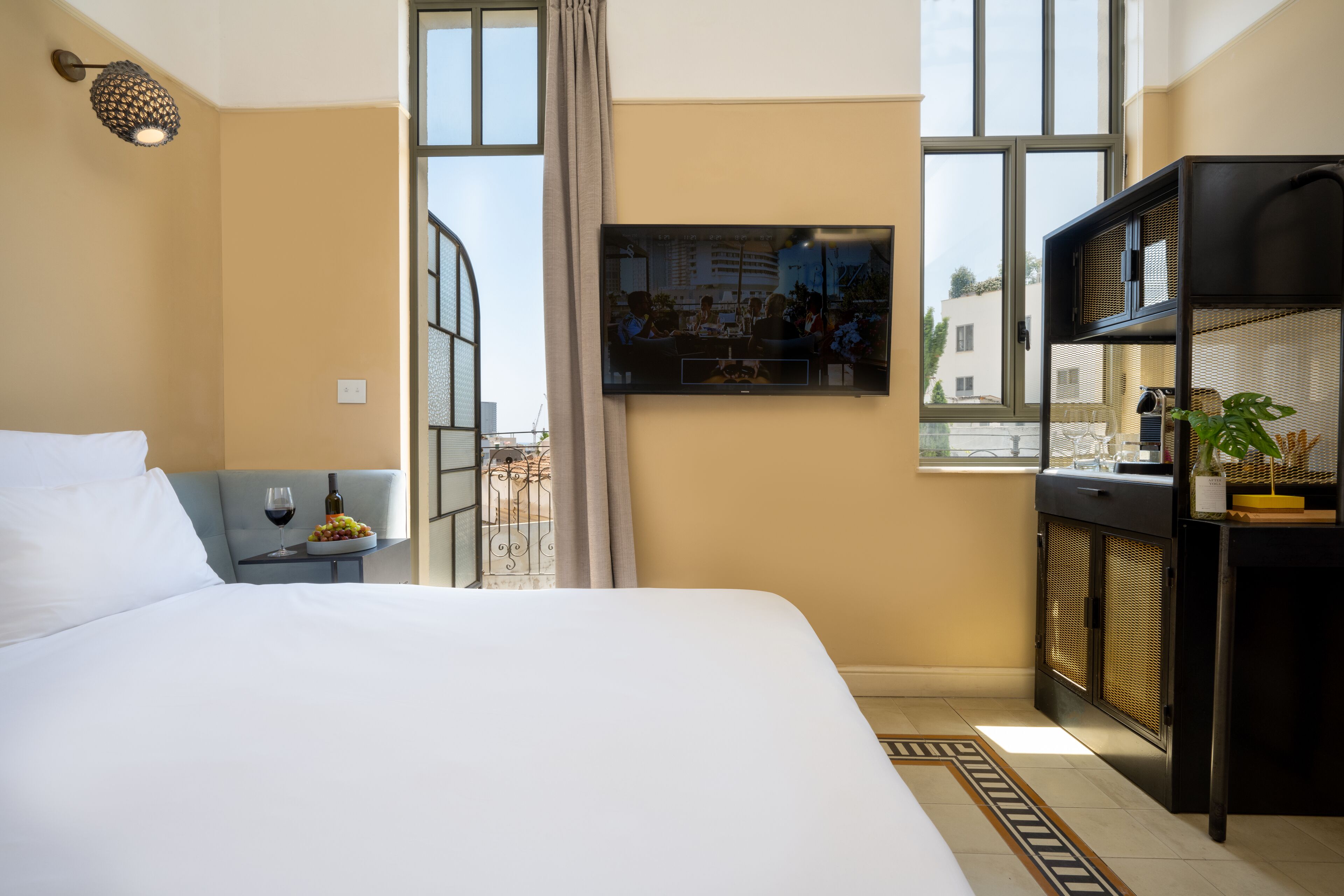 A hotel room with a bed, a small table with a wine glass and fruit, a TV on the wall, and a view of the city through large windows.