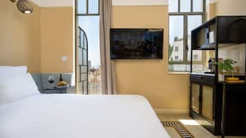 A hotel room with a bed, a small table with a wine glass and fruit, a TV on the wall, and a view of the city through large windows.