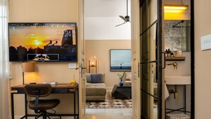 A hotel room with a bed, a desk with a chair, a TV displaying a sunset, a sofa, a small table with a lamp, and a bathroom with a mirror and sink. - Assemblage Boutique (Tel Aviv)