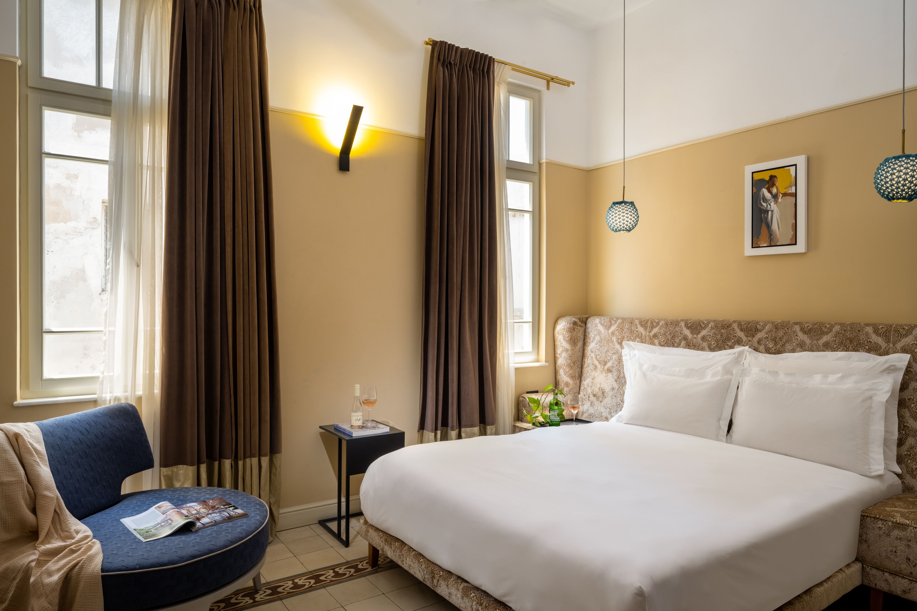 A hotel room with a large bed, a blue armchair, a small table with a magazine and a bottle of wine, and a painting on the wall.
