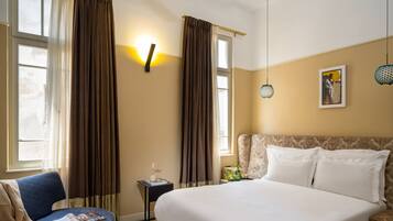 Une chambre d’hôtel avec un grand lit, un fauteuil bleu, une petite table avec un magazine et une bouteille de vin, et un tableau accroché au mur.