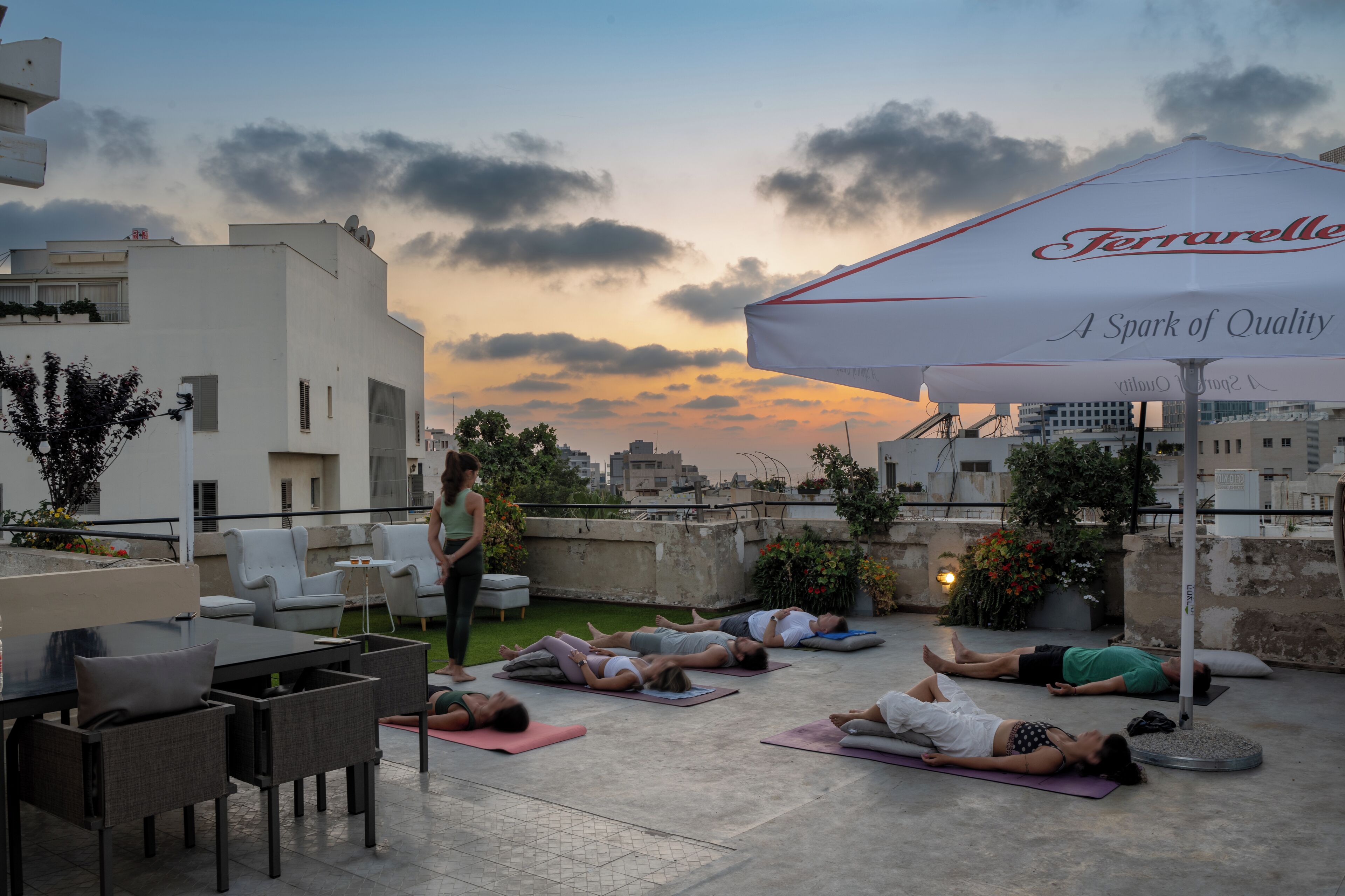 Yoga-Kurs auf einem Dach mit Teilnehmern auf Yogamatten, einem weißen Sonnenschirm und Stadtgebäuden im Hintergrund.