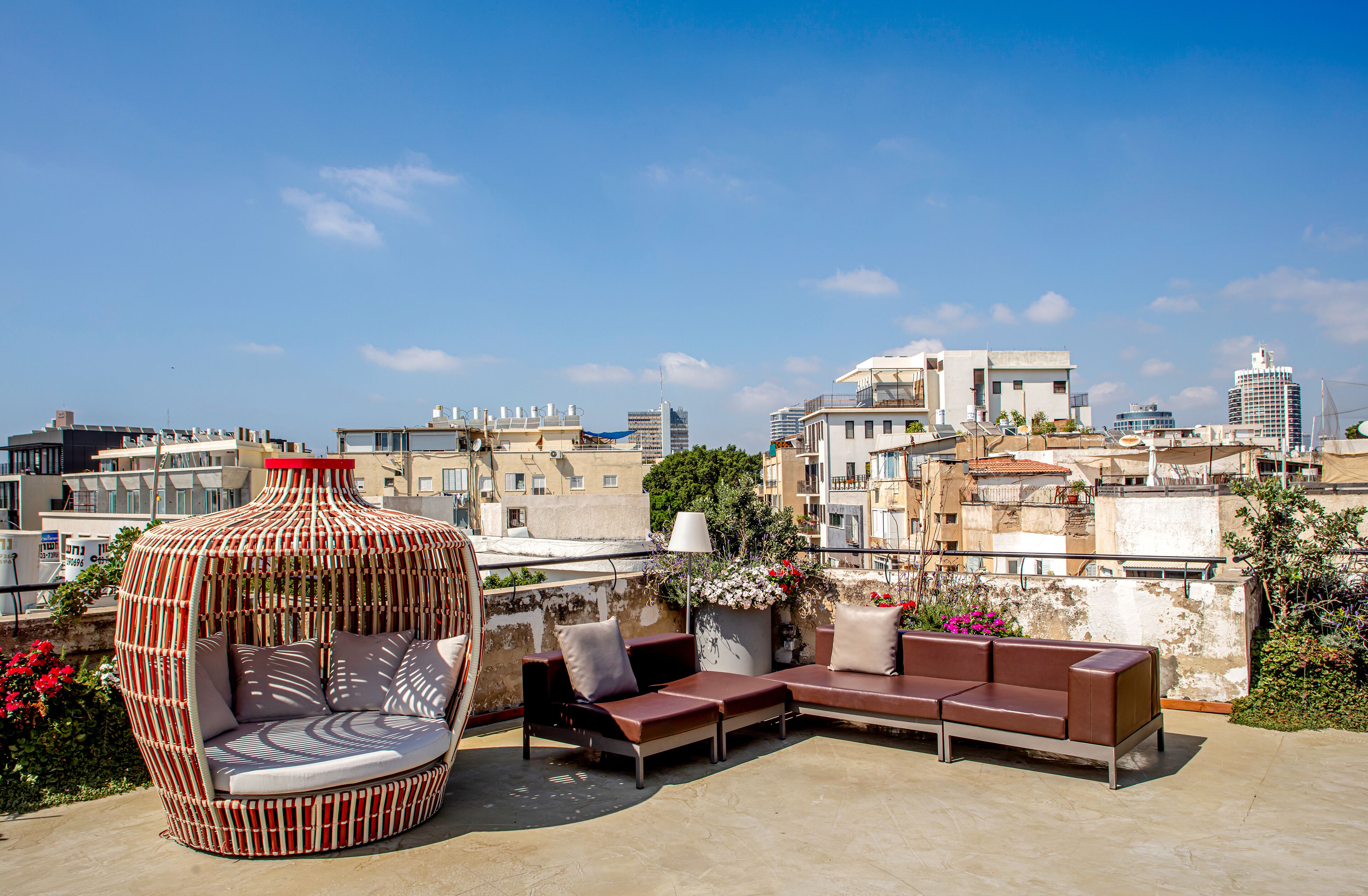 A rooftop terrace with a wicker chair, a sofa, and a table, overlooking a cityscape with buildings and a clear sky.