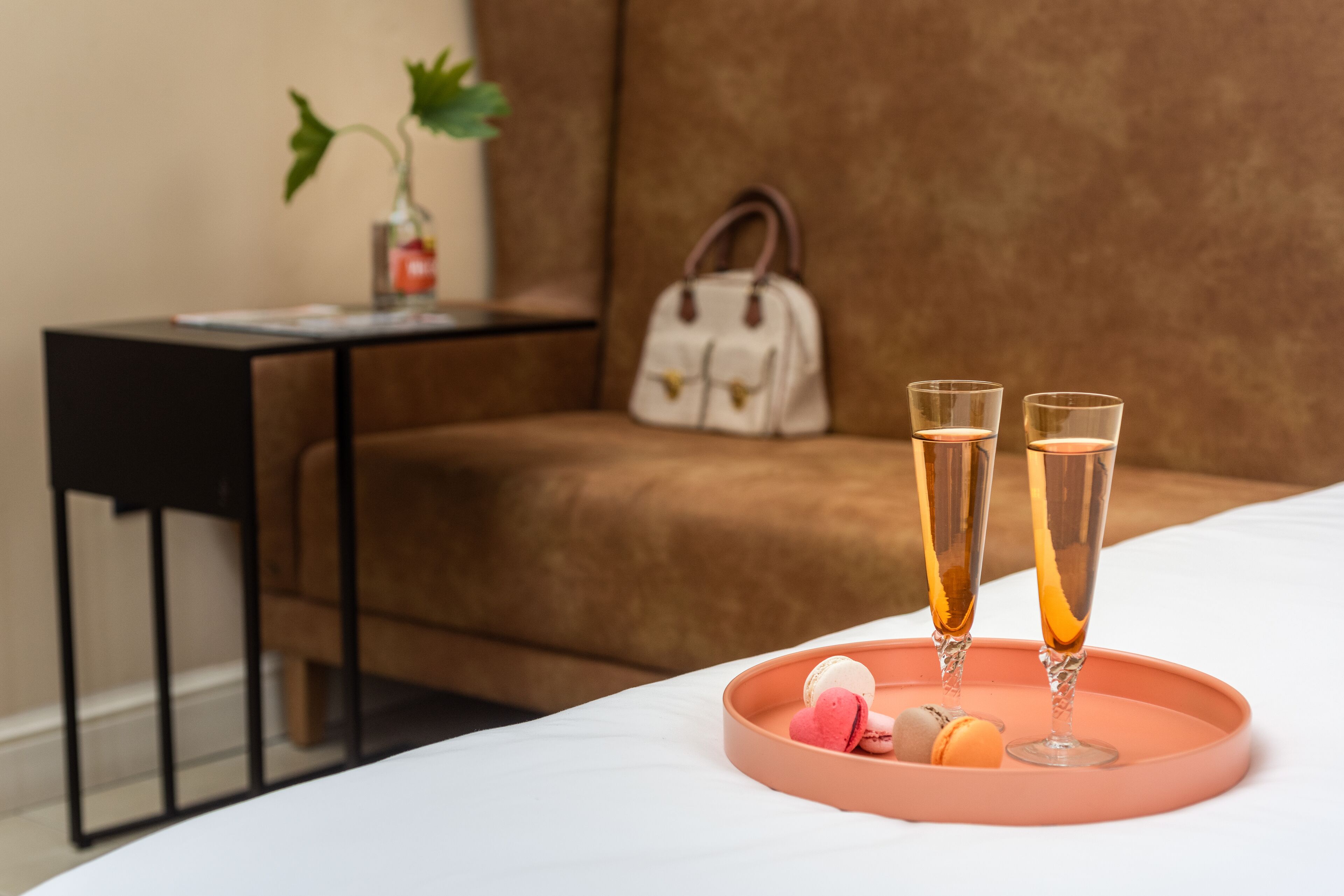 A bed with a white bedspread, a pink tray with two glasses of champagne and macarons, a brown sofa, a small table with a plant, and a white handbag.