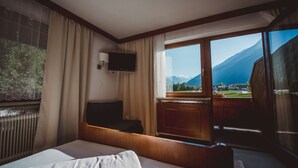 A hotel room with a bed, a TV mounted on the wall, and a large window offering a mountain view. - Alpenhaus Monte (Neustift Im Stubaital)