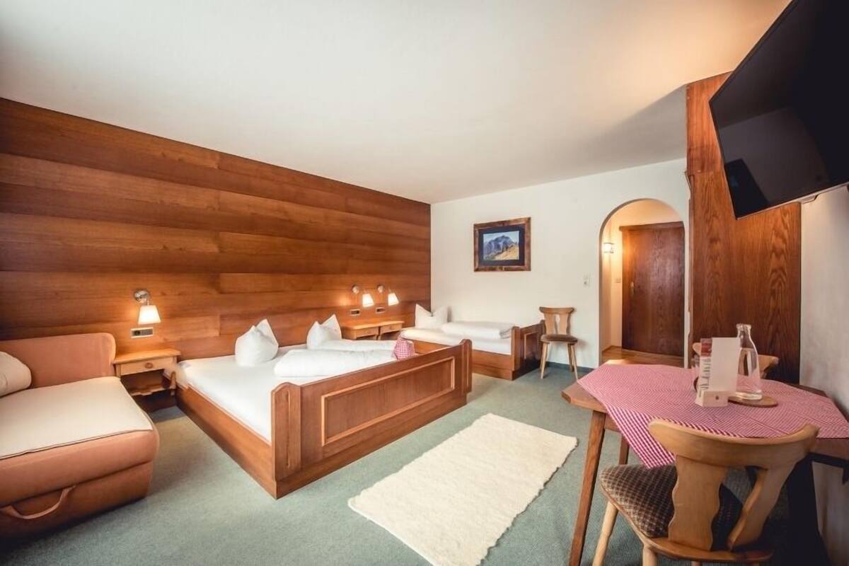 A hotel room with a wooden headboard bed, a small desk with a chair, a table with a red and white checkered tablecloth, and a framed picture on the wall.