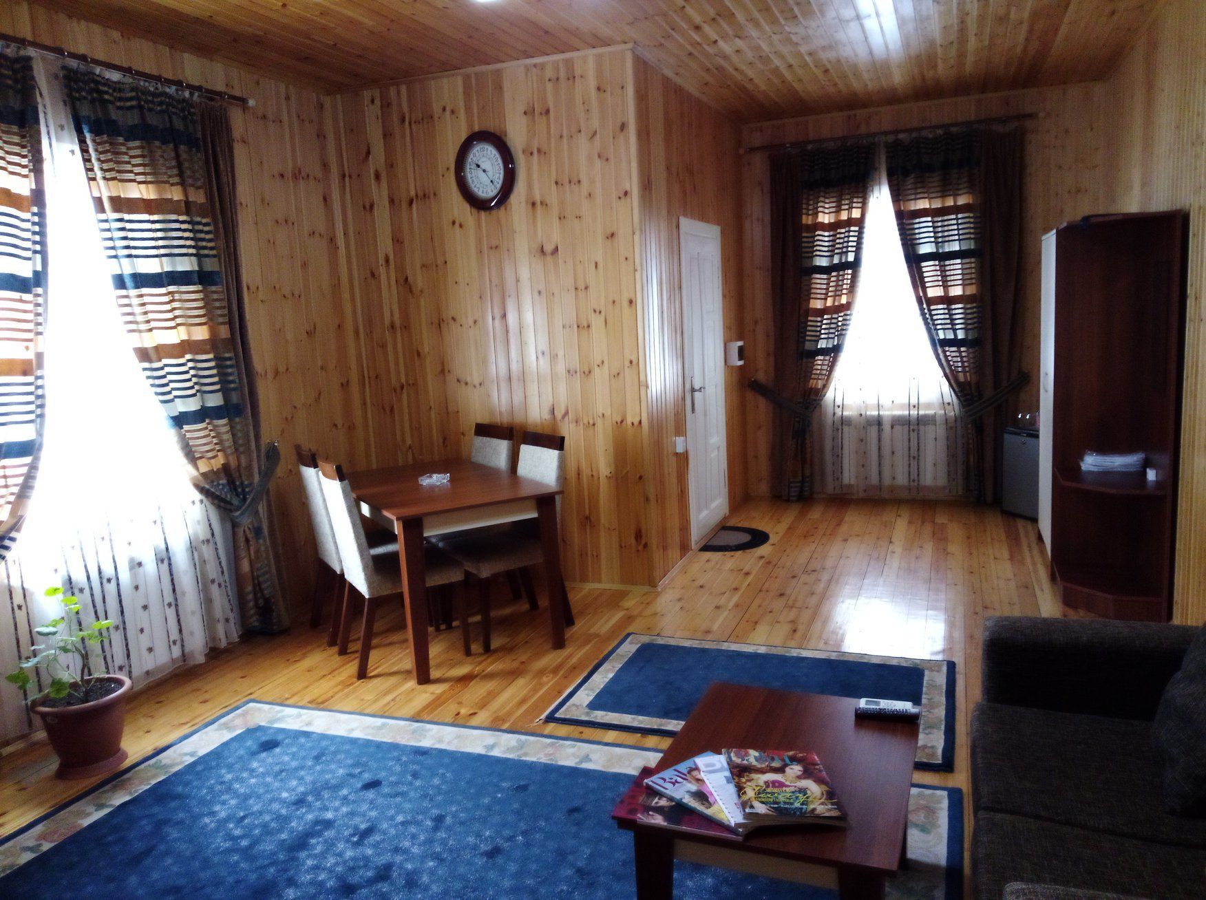 A wooden interior with a dining table, chairs, a sofa, a bookshelf, and a clock on the wall.