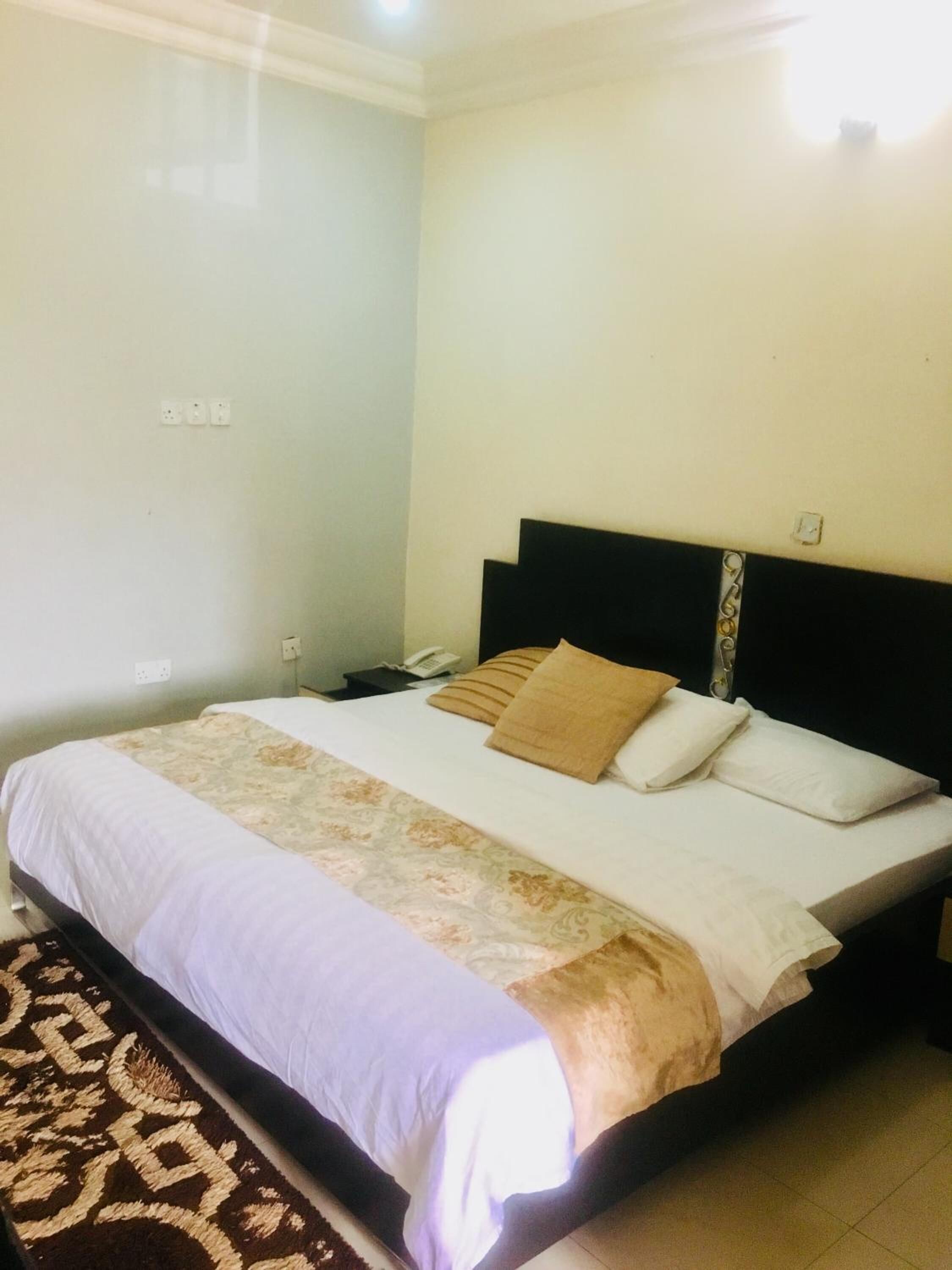 A neatly made bed with a patterned bedspread and two pillows, a bedside table with a phone, and a wall socket.