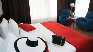 Et hotelværelse med en stor seng, to blå lænestole, et lille bord med en lampe, og udsigt udad gennem store vinduer.