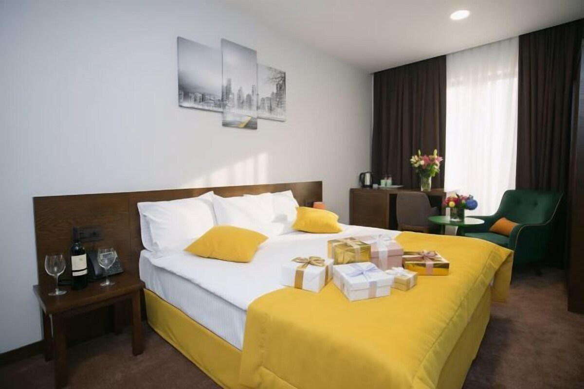 A hotel room with a large bed, a bedside table with a bottle and glasses, a chair, a small table with a vase of flowers, and two wall art pieces.