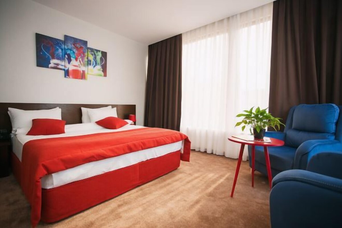 A hotel room with a large bed, a blue armchair, a small table with a plant, and two abstract paintings on the wall.