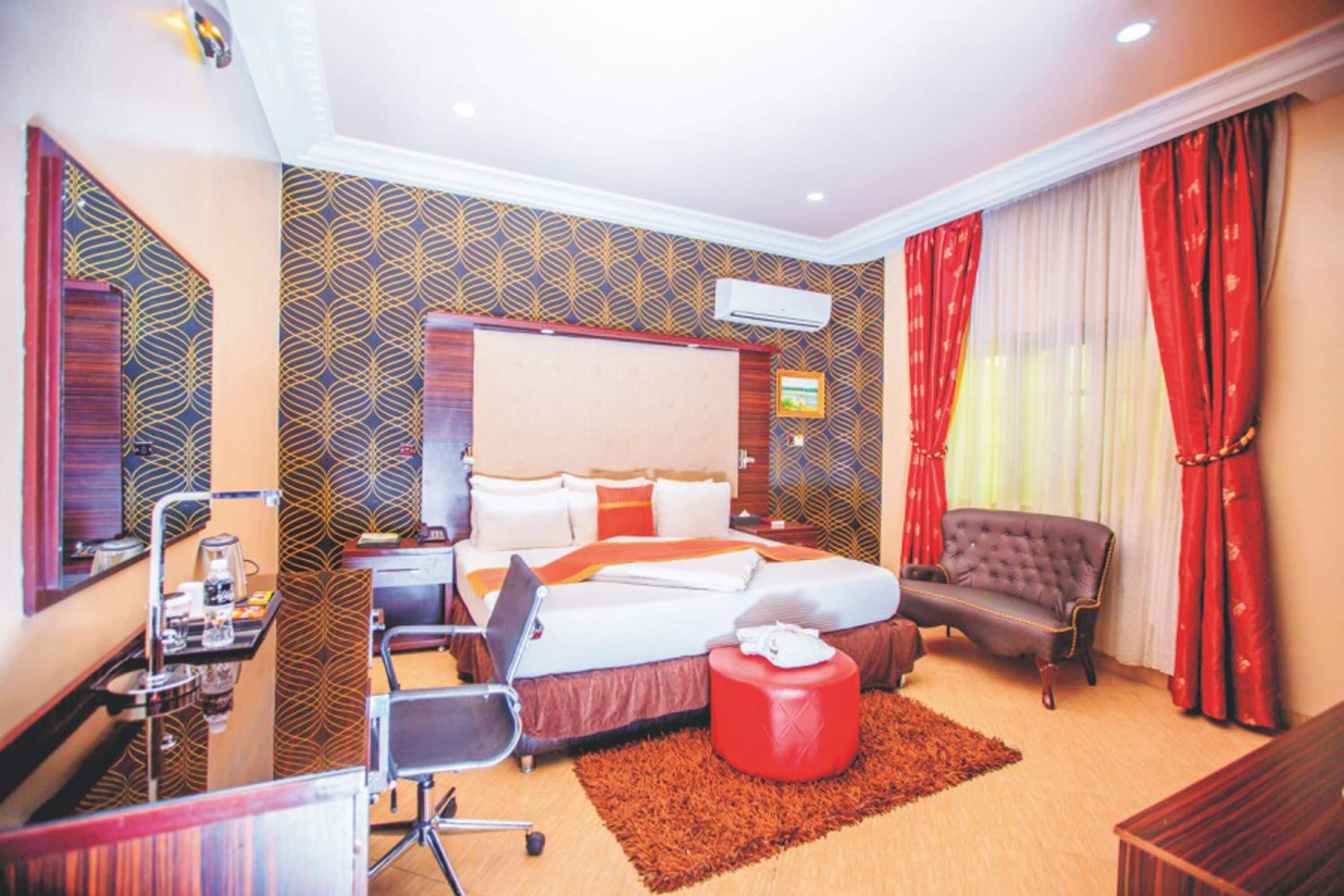 A hotel room with a bed, a desk with a chair, a red ottoman, a red armchair, and a red curtain.