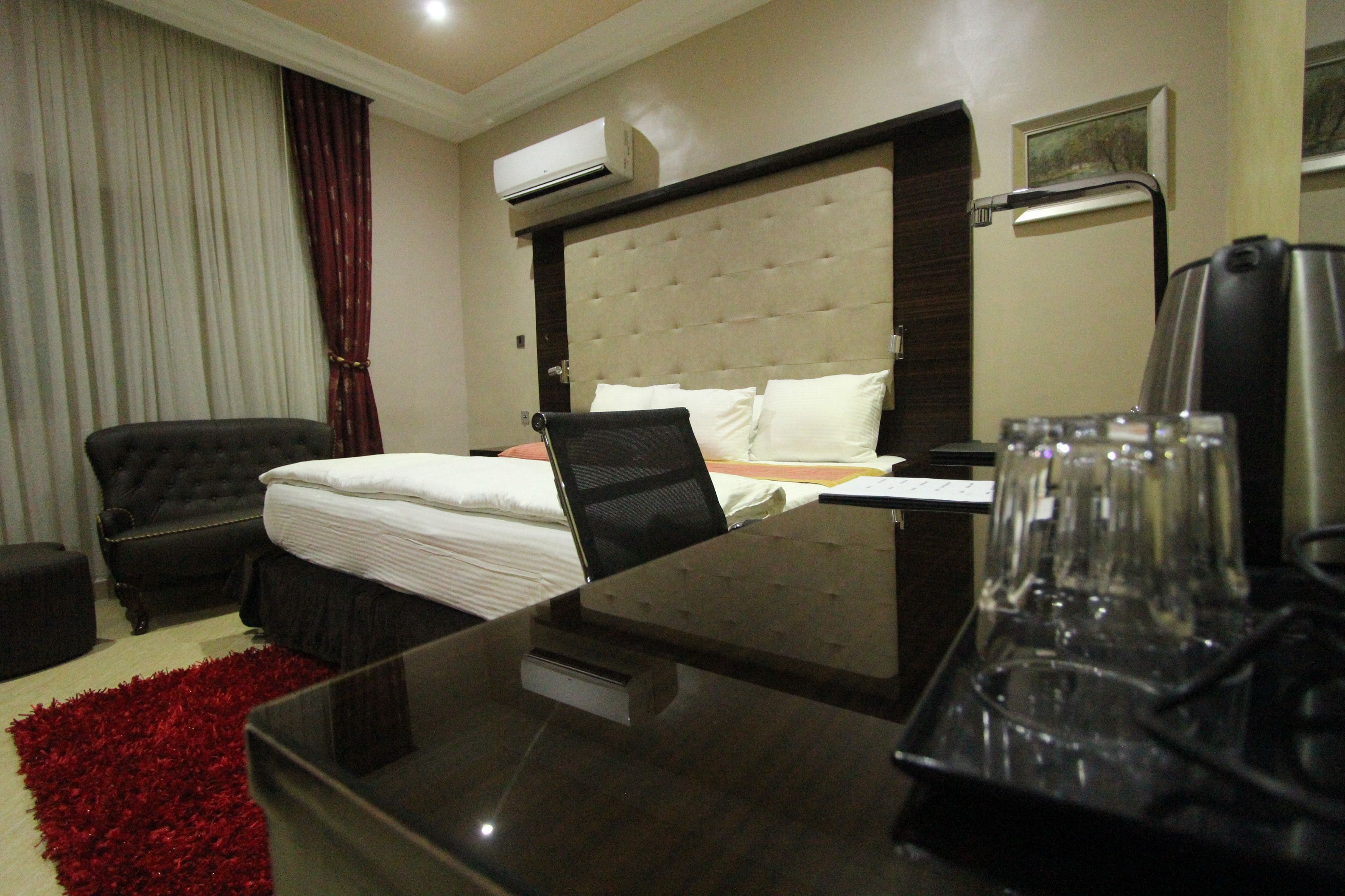 A hotel room with a bed, a desk with a laptop, a chair, a red rug, an air conditioner, and a painting on the wall.