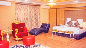 A hotel room with a large bed, a red armchair, a black lounge chair, a small table with a glass, and a chandelier. - Eterno Hotels Limited (Benin City)