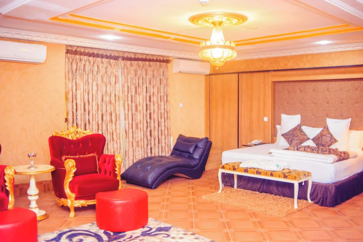 Une chambre d’hôtel avec un grand lit, un fauteuil rouge, une chaise longue noire, une petite table avec un verre et un lustre.