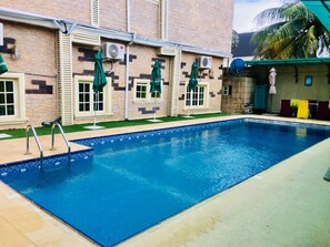 A swimming pool with a tiled edge, surrounded by a brick building with shutters, and a palm tree in the background.