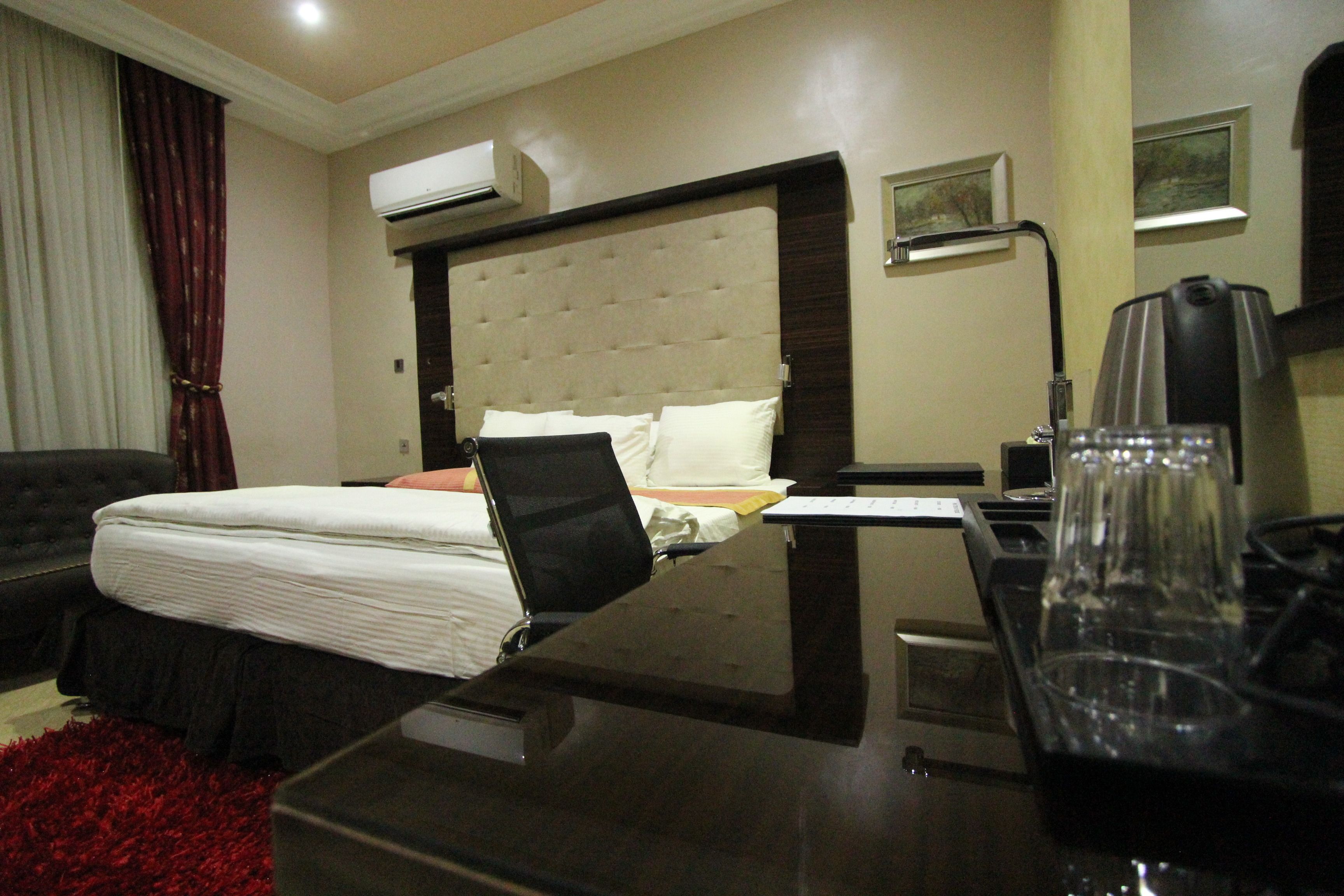 A hotel room with a bed, a desk, a chair, a red rug, an air conditioner, and a lamp.