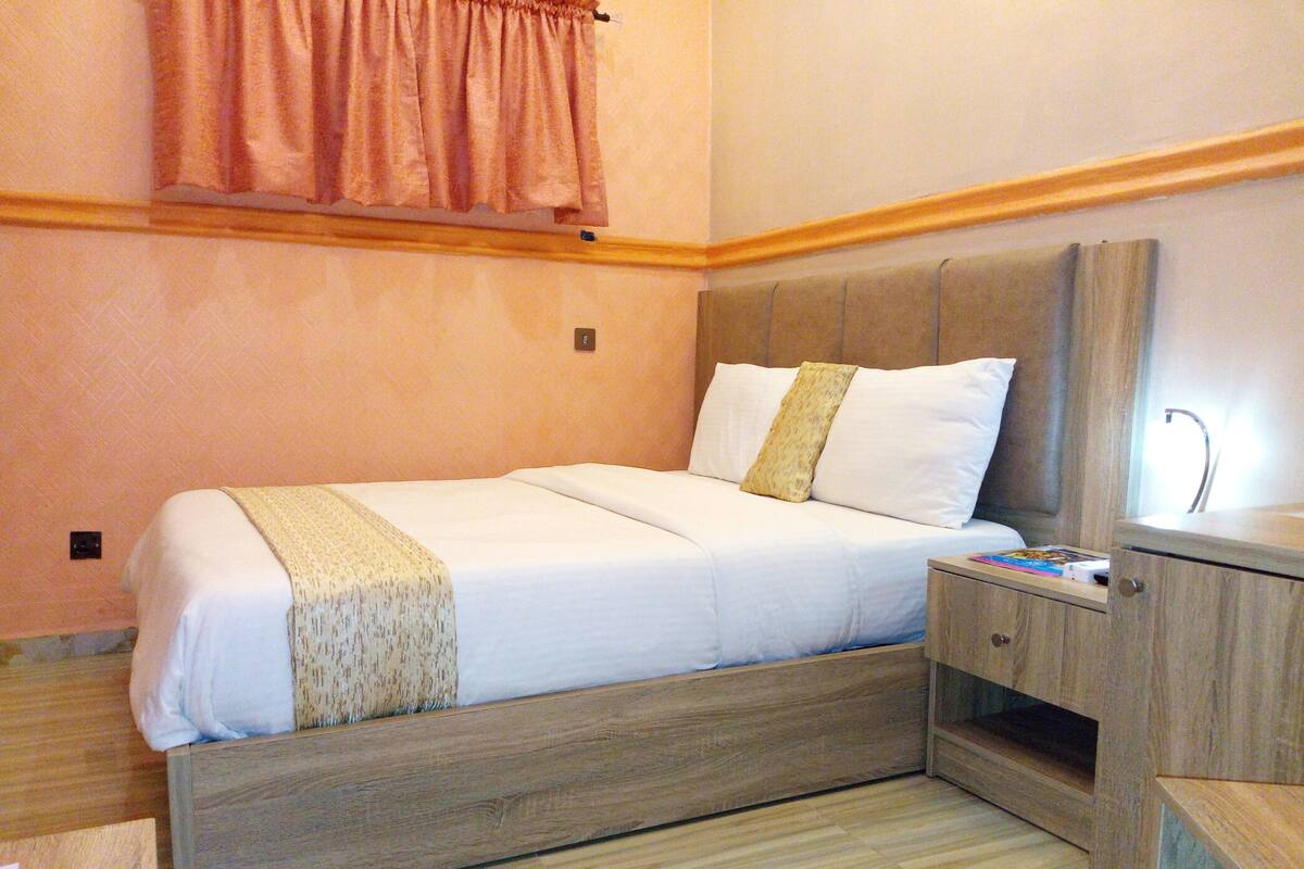 A neatly made bed with a wooden headboard, a bedside table with a book, and a small wooden cabinet.