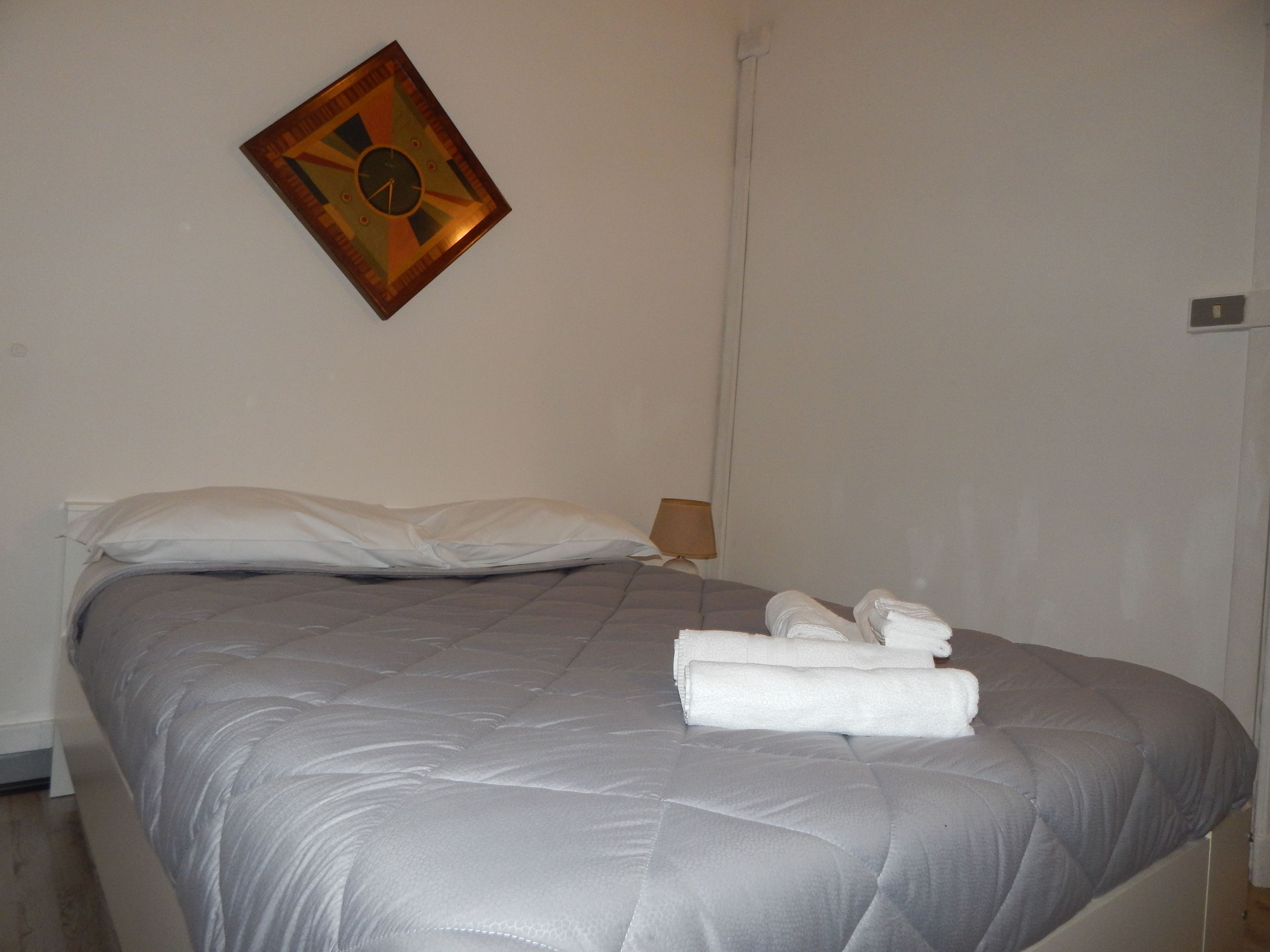 Una cama bien hecha con colcha gris, almohadas blancas y toallas dobladas al lado.