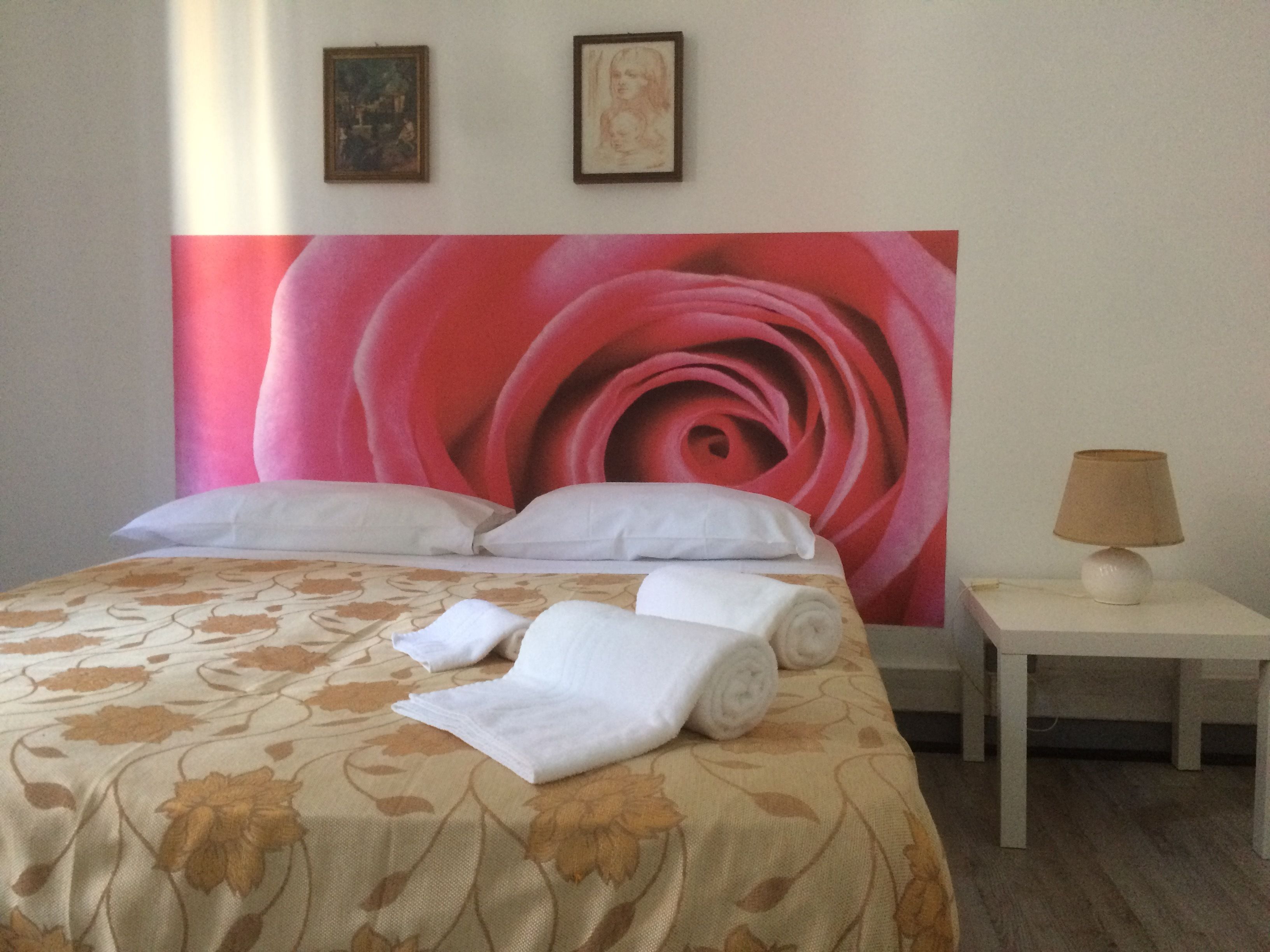 A hotel room with a large floral bedspread, a neatly made bed with white linens, a nightstand with a lamp, and two framed pictures on the wall.
