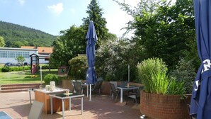 Een terras buiten met een tafel, stoelen, een blauwe parasol en een bak met planten van baksteen.