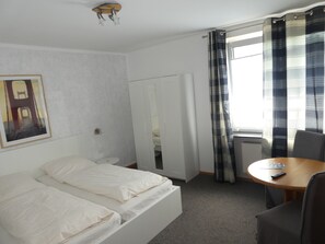 A hotel room with a bed, a small table, a chair, a window with curtains, a radiator, and a painting on the wall.