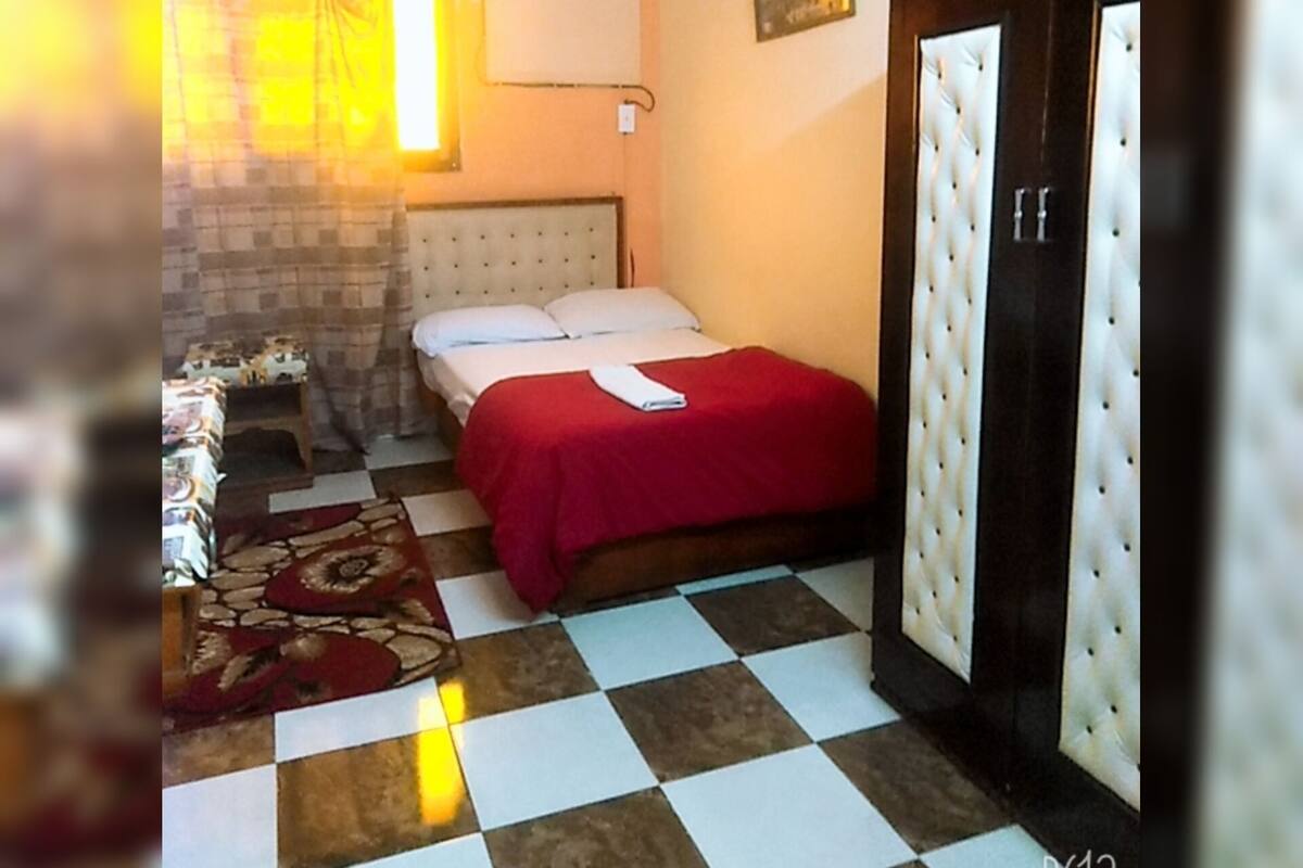 A bedroom with a checkered floor, a bed with a red bedspread, a wooden wardrobe, and a window with curtains.