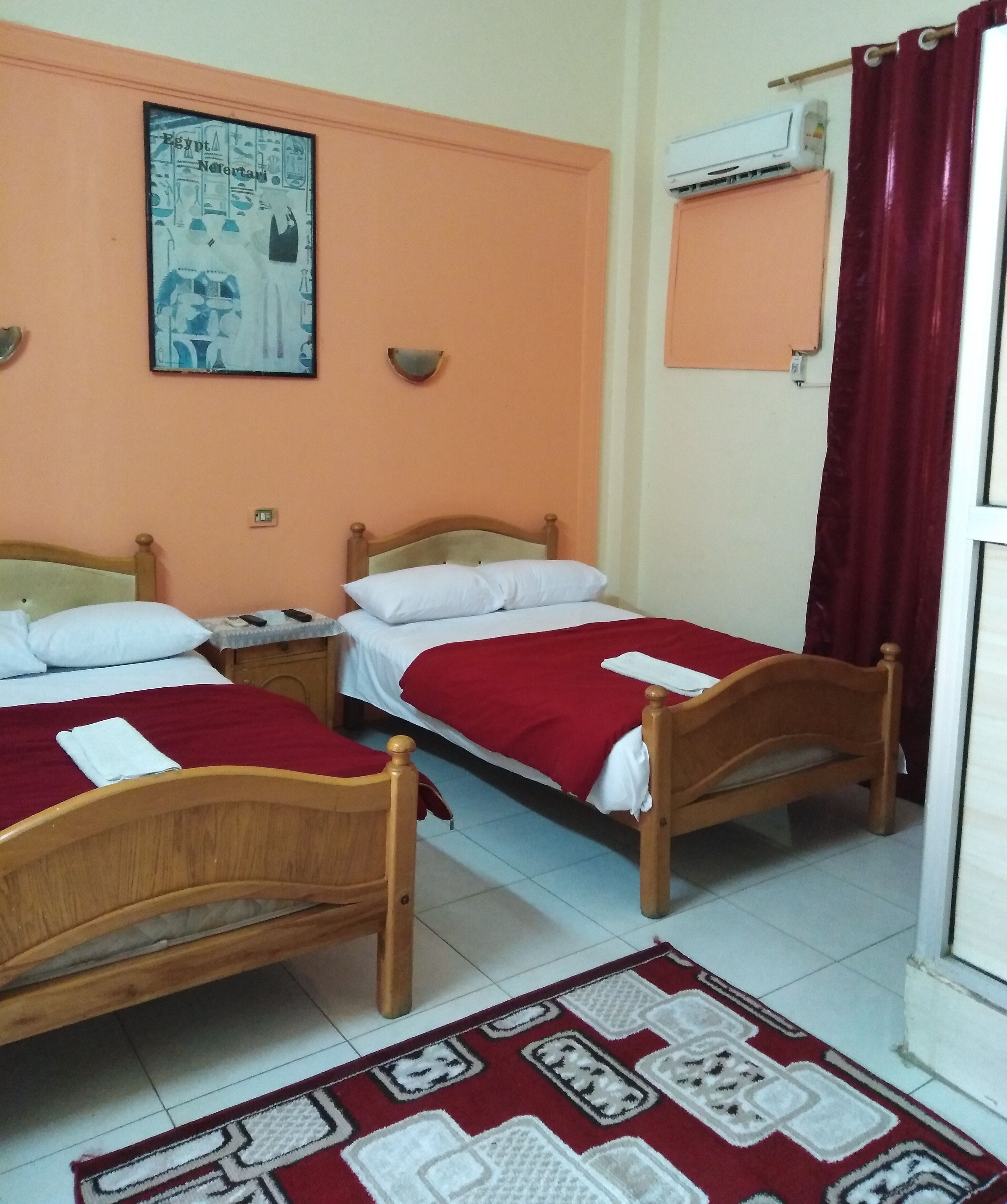 A hotel room with two wooden beds, a red and white patterned rug, a wall-mounted air conditioner, and a painting of Egyptian hieroglyphs.