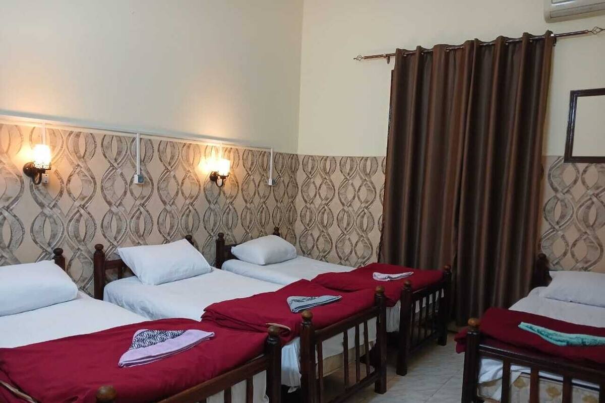 A hotel room with four single beds, each with a wooden frame and white bedding. There are two wall-mounted light fixtures on the left side of the room.