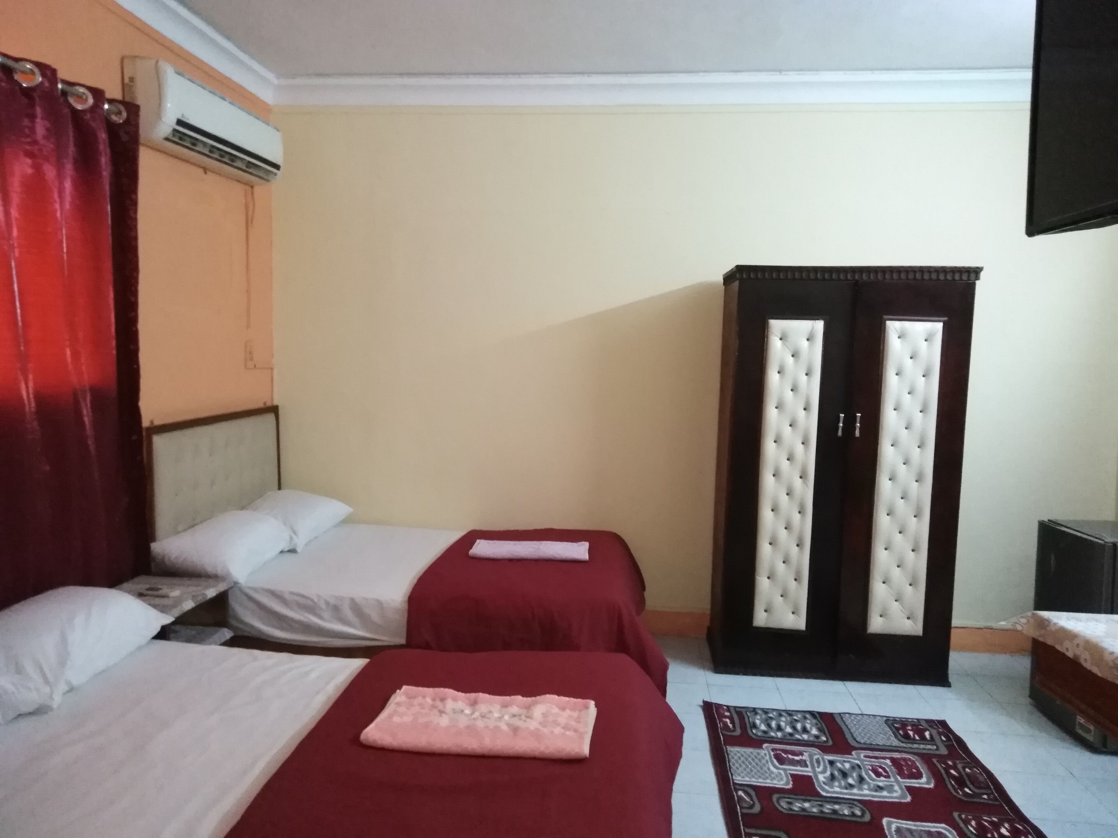 A hotel room with two beds, a red curtain, an air conditioning unit, a wardrobe with a mirror, and a patterned rug.