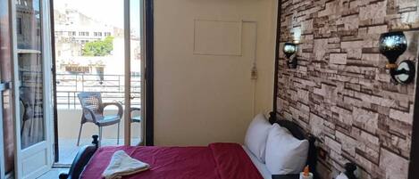 A hotel room with two beds, a balcony with chairs, an air conditioning unit, and a brick-patterned wall.