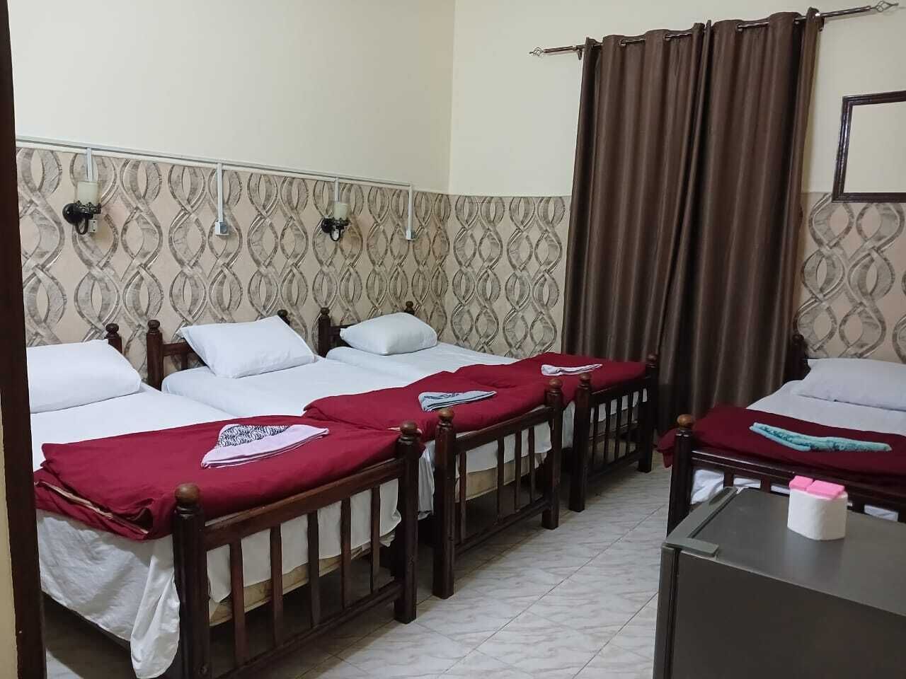 Une chambre d’hôtel avec deux lits, chacun doté d’un cadre en bois et de draps blancs. On y trouve une petite table sur laquelle est posée une boîte de mouchoirs, et un rideau est partiellement tiré sur le côté.