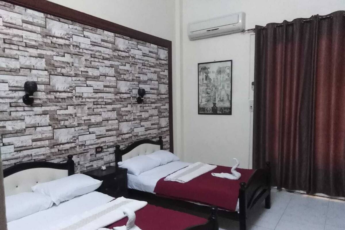A hotel room with two beds, a stone-patterned wall, an air conditioning unit, a framed picture, and a curtain.