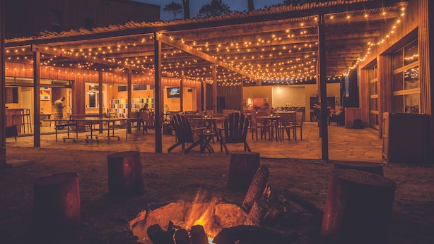 Un'area patio all'aperto con luci a stringa, tavoli e sedie in legno e un braciere.