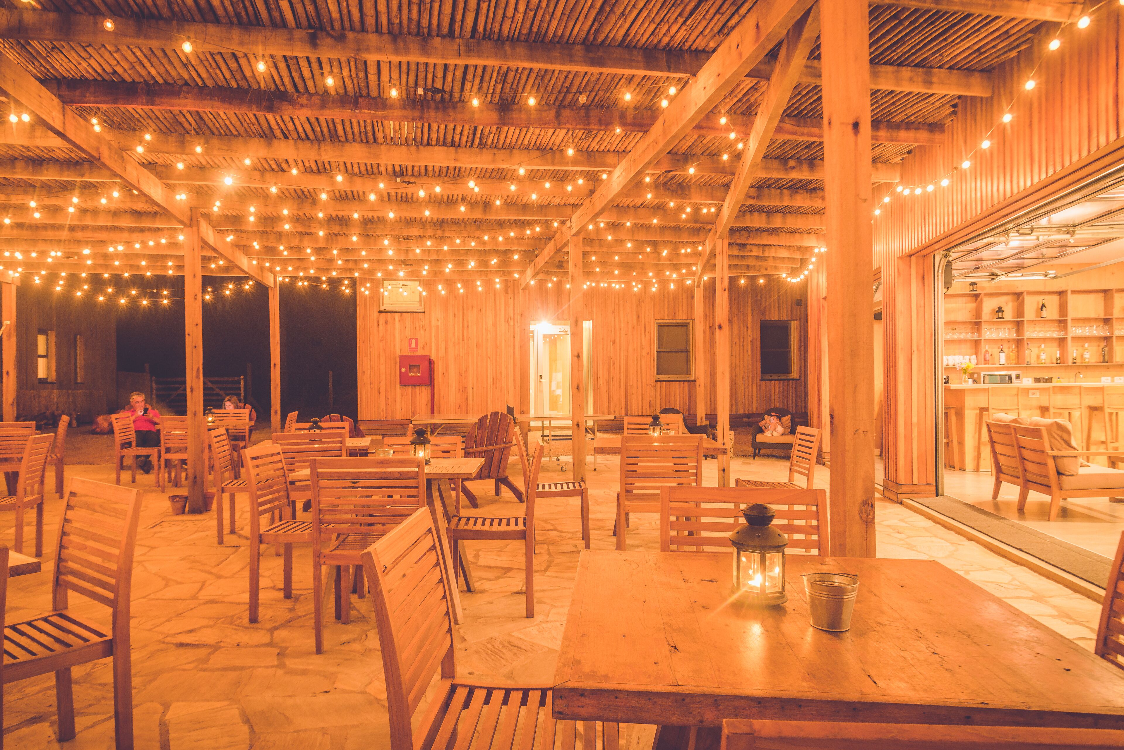 A wooden interior with tables and chairs, string lights, and a visible kitchen area.