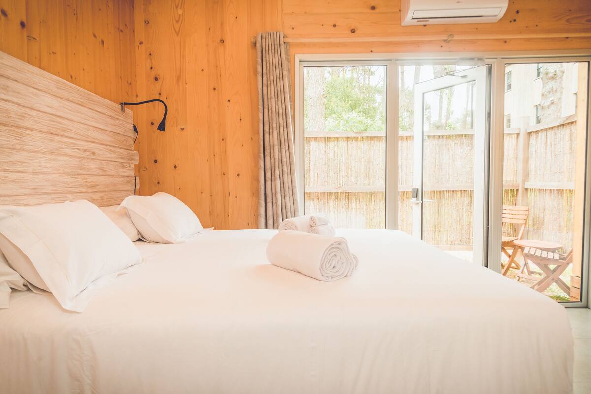 Camera da letto con un ampio letto, lenzuola bianche, una testiera in legno e una porta scorrevole in vetro che dà su una zona esterna con recinzione in legno.