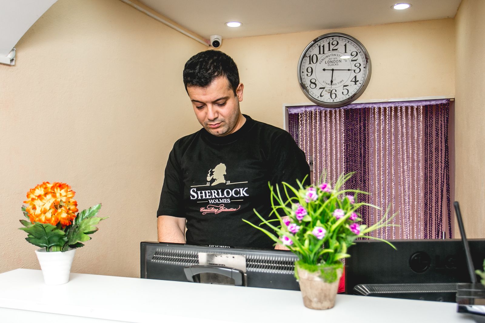 A person standing behind a desk with a computer, a clock on the wall, and two potted plants.